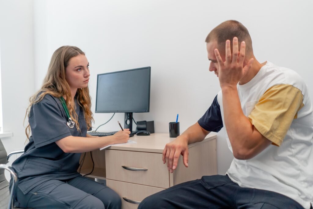 Young man holding his head while female clinician listens and takes notes during consultation.