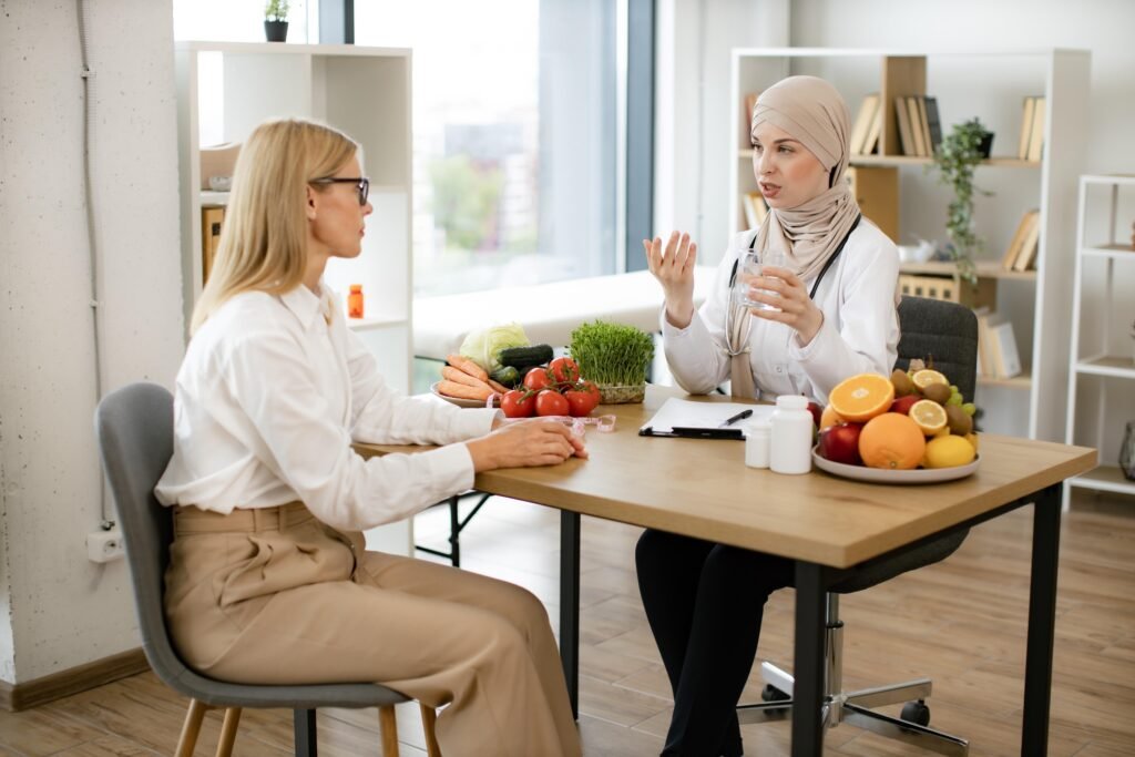 Dietitian in hijab advising woman about nutrition at a desk with fruits and vegetables.