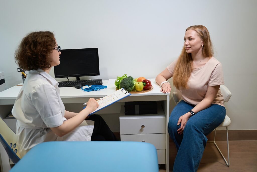 Dietitian taking notes while talking with woman during nutrition consultation, plate of fresh vegetables on desk between them.